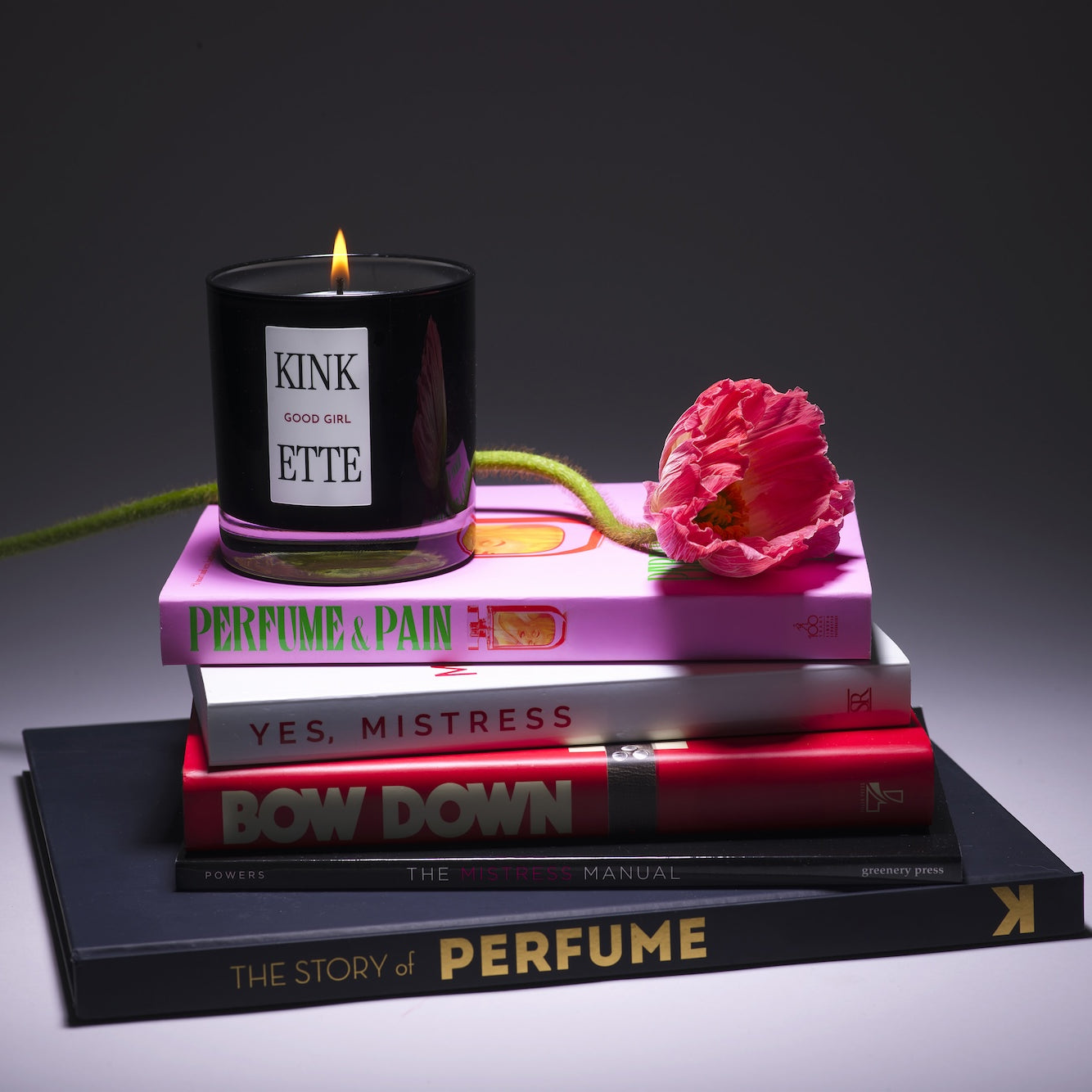Kinkette GOOD GIRL luxury candle on stack of books including Perfume & Pain and Yes Mistress with pink poppy