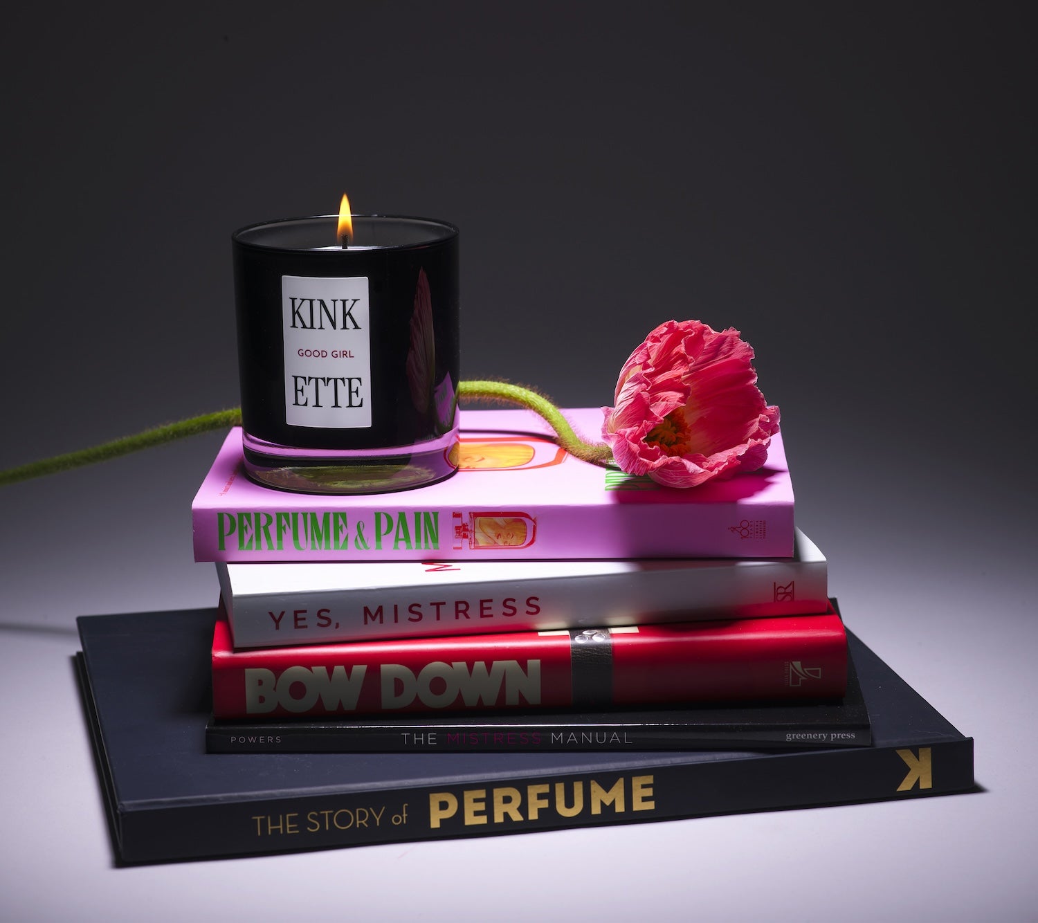 Kinkette GOOD GIRL luxury candle on stack of books including Perfume & Pain and Yes Mistress with pink poppy