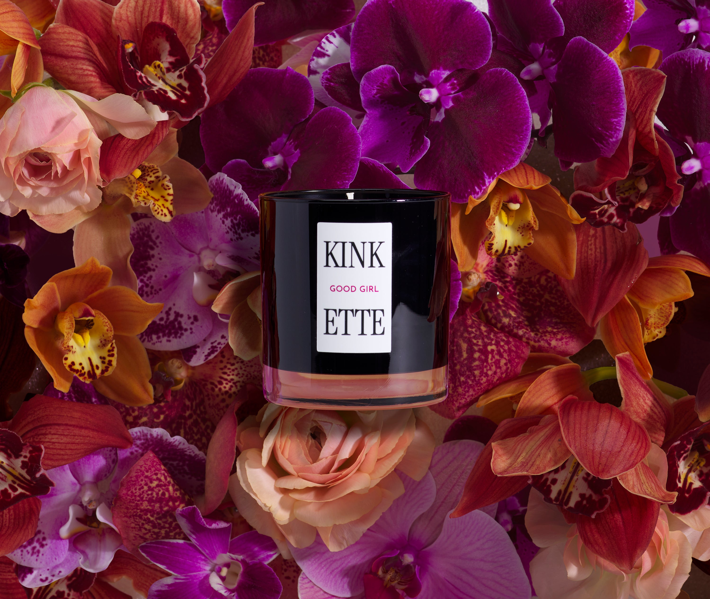 Kinkette GOOD GIRL Signature Candle surrounded by purple orchids and warm orange flowers — luxury kink-inspired fragrance