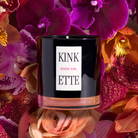 Black candle labeled 'KINKETTE GOOD GIRL' surrounded by pink and orange flowers