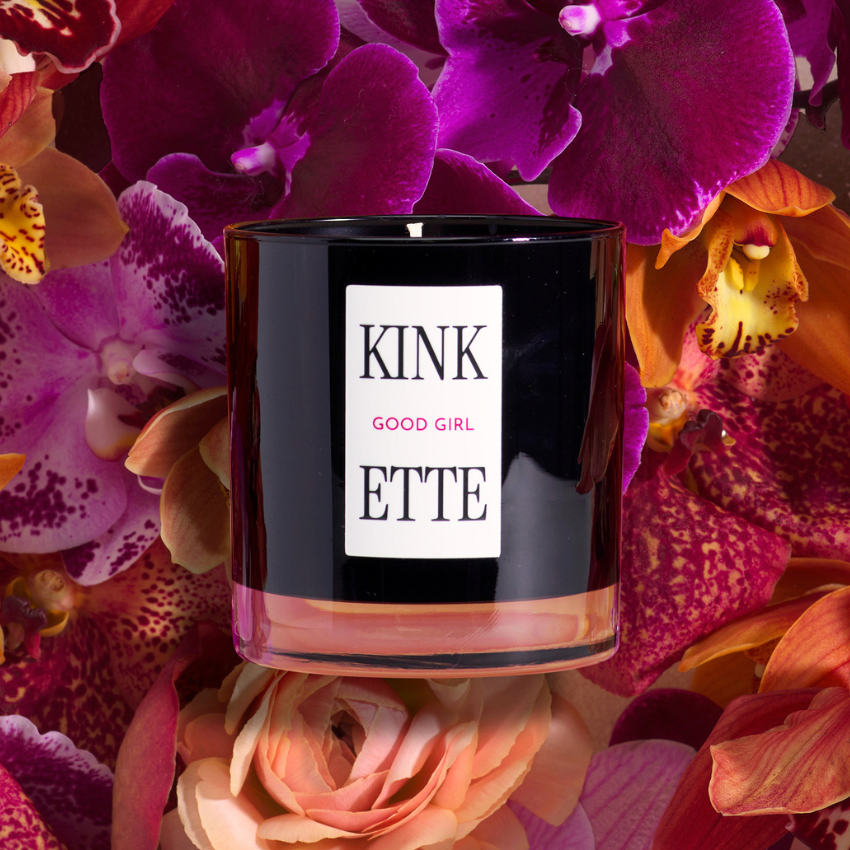 Black candle labeled 'KINKETTE GOOD GIRL' surrounded by pink and orange flowers