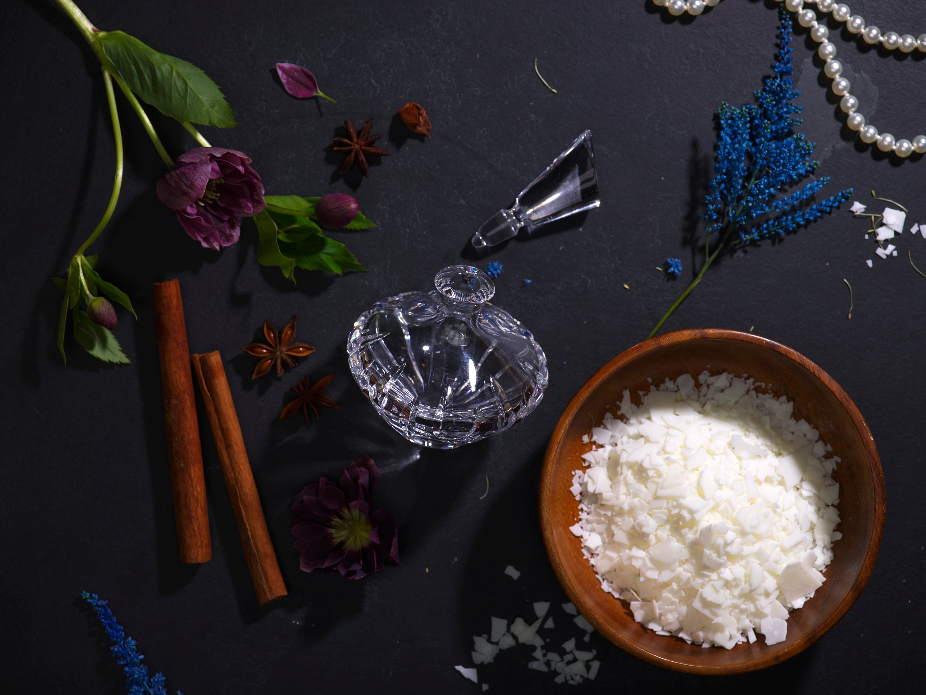 Kinkette Scent Philosophy — coconut soy wax flakes, flowers, cinnamon, and crystal perfume bottle on dark surface